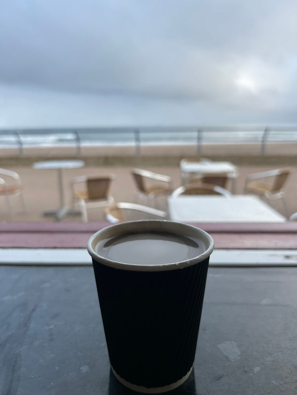Close-up of a disposable cup of coffee sitting on a dark surface. The background is out of focus but clearly depicts an outdoor cafe setting overlooking a beach on an overcast day. Several empty tables and chairs are visible. The overall mood is tranquil and slightly melancholic.