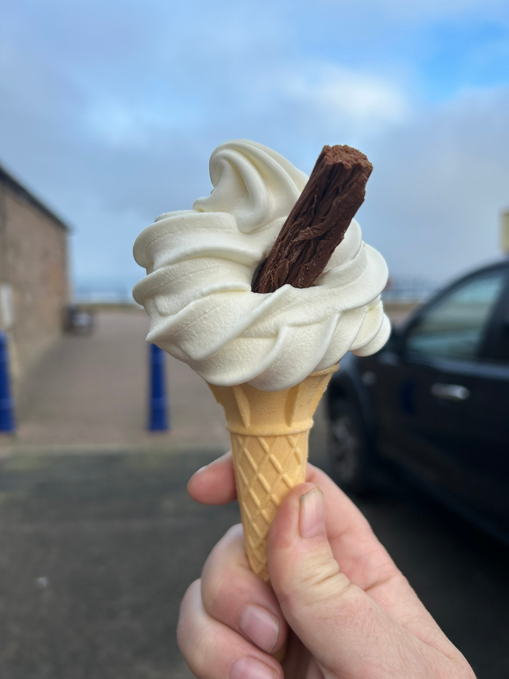 Close-up of Leonie's hand holding a soft-serve ice cream cone. The ice cream is white and has a chocolate flake on top. The background is blurred but shows a seaside location with a building and a car.
