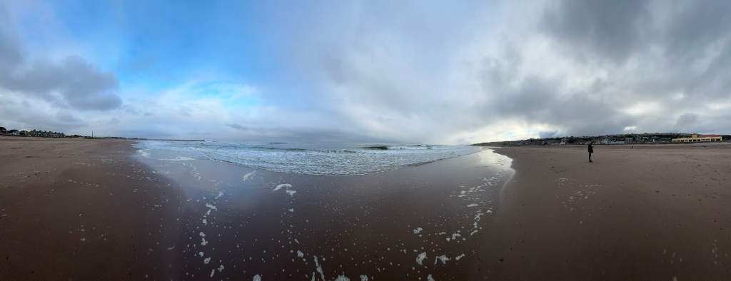 Panoramic view of a wide, sandy beach with gentle waves lapping the shore. The sky is mostly cloudy, with patches of blue visible. A single, small figure is visible in the distance walking along the beach. Buildings are visible in the far distance on either side of the beach. The overall mood is serene and somewhat melancholic, due to the overcast sky and the solitary figure.