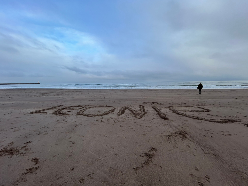 Charlie walking away from the viewer on a beach. The sand in the foreground has the name Leonie written in large letters. The ocean is calm with a mostly overcast sky. A breakwater is visible in the distance. The overall mood is serene and slightly melancholic.