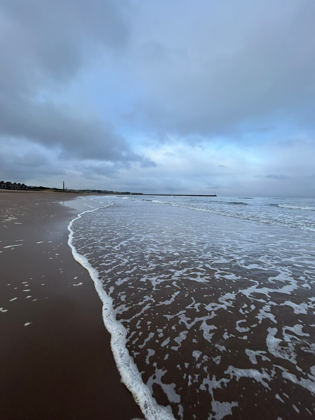 Each scene under a cloudy sky. The ocean is calm with small waves gently lapping onto the dark sand. A long, low structure, possibly a breakwater, is visible in the distance along the shoreline. The overall mood is serene and somewhat melancholic due to the muted colours and overcast sky.