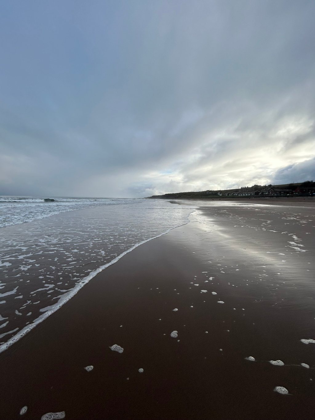 Wide, sandy beach under a cloudy sky. Gentle waves lap the shore, leaving a trail of white foam on the dark sand. In the distance, a line of buildings is visible along the coast. The overall mood is serene and somewhat melancholic due to the overcast conditions.