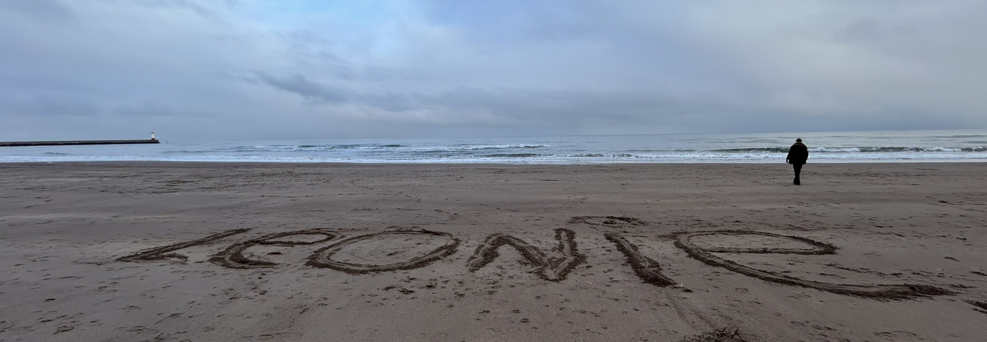 Charlie walking away from the viewer on a beach towards the ocean. The name Leonie is written in the sand in large letters near the foreground. The sky is overcast and the overall atmosphere appears sombre or reflective. A breakwater and lighthouse are visible in the distance.