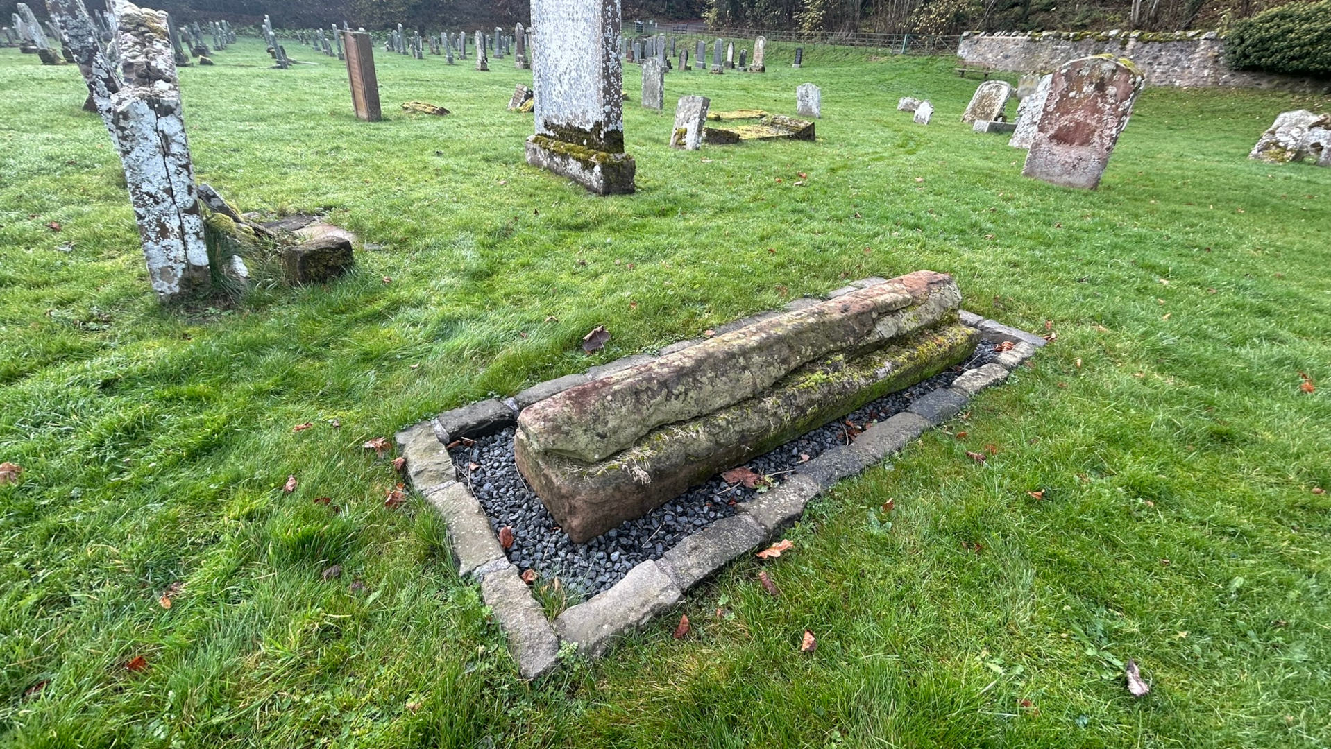Main focus is a long, rectangular stone, possibly a grave marker or a piece of a larger monument, set within a low stone border filled with dark gravel. This is situated in a grassy area amidst numerous other, more traditional, gravestones and markers, some of which are broken or dilapidated. The overall impression is one of age and decay within a rural or historical setting.