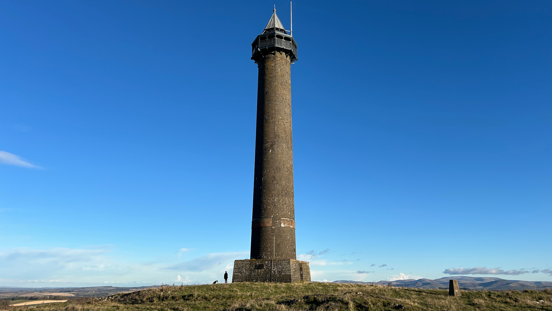 A  tall, slender stone tower standing atop a grassy hill against a clear, vibrant blue sky. A single person stands near the base of the tower, providing a sense of scale. The landscape is relatively flat, extending to a distant horizon. The overall impression is one of serenity and openness.
