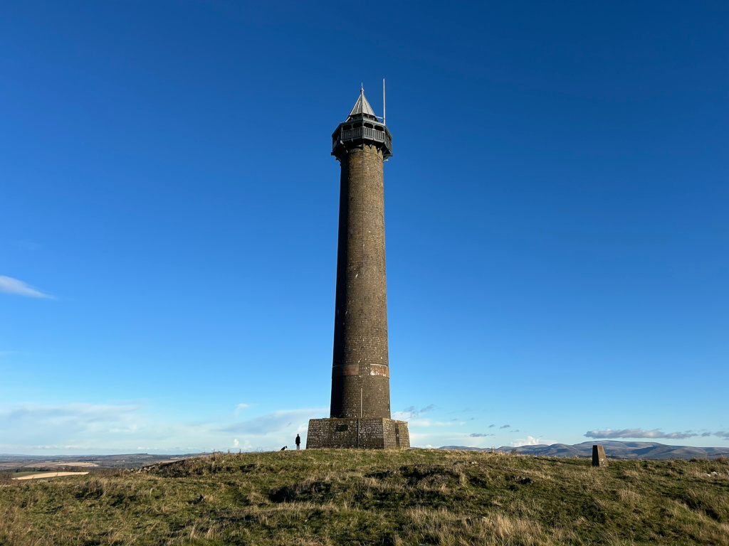 Tall, slender stone tower standing atop a grassy hill against a clear, vibrant blue sky. A single person stands near the base of the tower, providing a sense of scale. The landscape is relatively flat, extending to a distant horizon. The overall impression is one of serenity and openness.