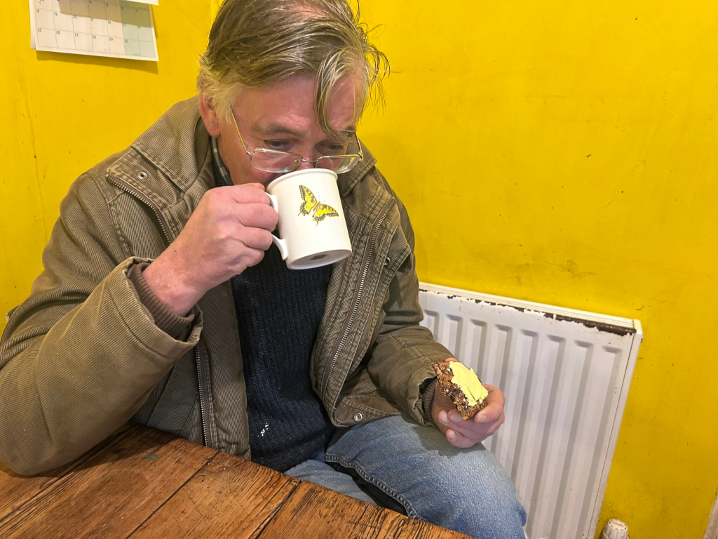Charlie with somewhat dishevelled light brown hair sitting at a wooden table. He’s wearing a corduroy jacket and a dark blue sweater. He's holding a mug with a yellow butterfly design, drinking from it, and simultaneously holding a piece of bread with yellow spread on it. The background is a vibrant yellow wall with a white radiator. The overall mood is simple and peaceful, suggesting a quiet moment of refreshment and possibly a solitary meal.