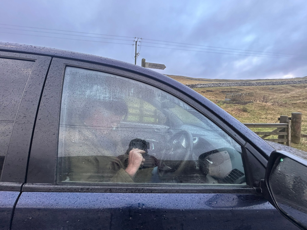 Charlie sitting inside a dark-blue car, partially obscured by rain-streaked windows. He appears to be holding a device, possibly a phone or camera, up to the window. The car is parked beside a rural road, with a dry-stone wall and a wooden gate visible in the background. The sky is overcast. The overall impression is one of quiet observation or documentation of the landscape.