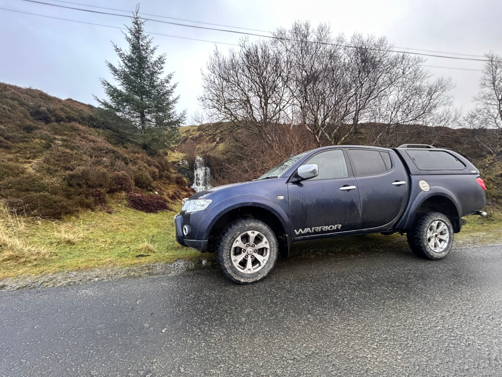 Dark-blue Mitsubishi L200 Warrior pickup truck parked on a wet road beside a small waterfall cascading down a hillside. A single evergreen tree stands prominently near the waterfall. The overall setting appears to be a remote, sparsely vegetated area, possibly in a mountainous or hilly region. The weather seems overcast.