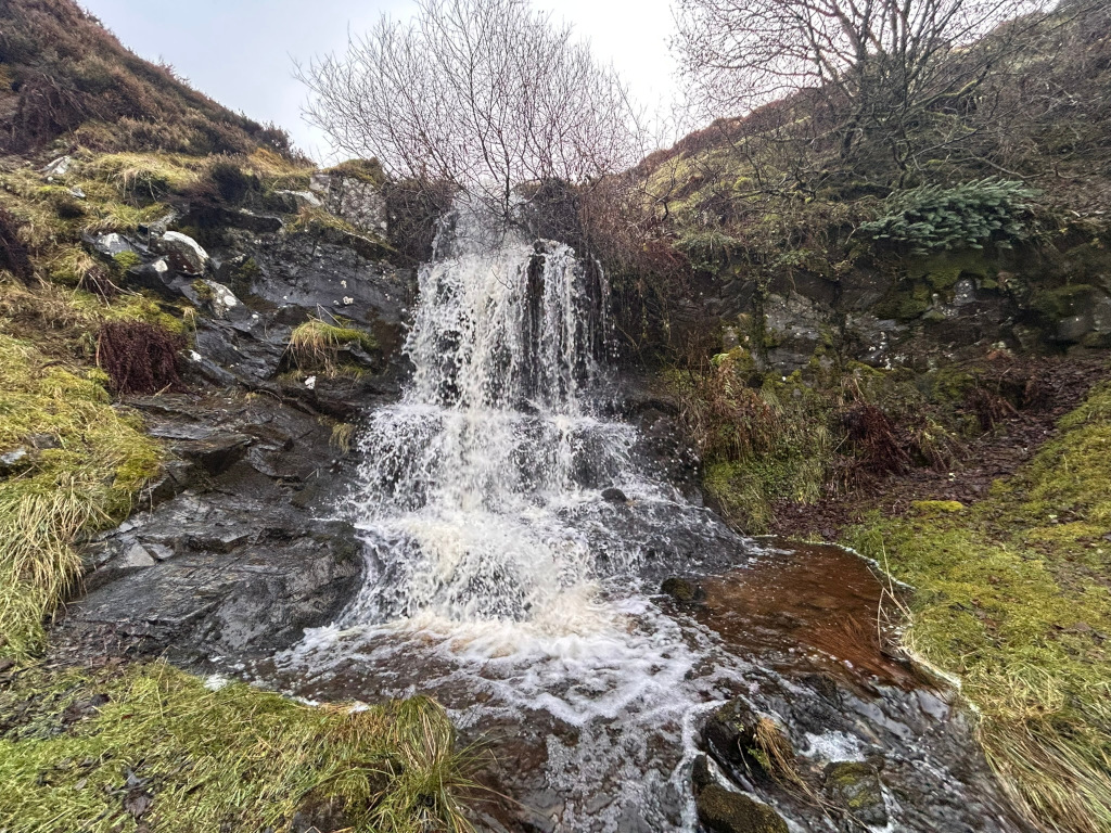 Small waterfall cascading down dark rocks, surrounded by lush green moss and vegetation. The water appears relatively clear and the overall setting is tranquil and natural, suggestive of a secluded or remote location. The sky is overcast.