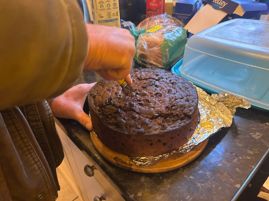 Charlie  using a knife to cut a slice of a dark fruitcake. The cake is round, sits on a wooden board, and is wrapped in aluminium foil. In the background are various food items, including bread and what appears to be other baked goods and packaging. The setting seems to be a kitchen counter. The overall impression is a domestic scene of cake preparation or serving.