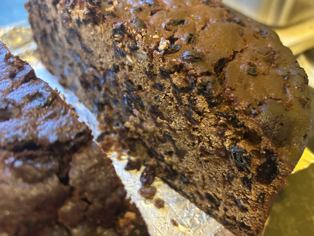 Close-up view of a sliced fruitcake. A large portion of the cake is visible, revealing its dense, dark brown texture and numerous dark-coloured fruits embedded throughout. A smaller, separate slice of the same cake is also in the picture. The cake rests on a piece of foil. The overall impression is one of a rich and possibly traditional baked good.
