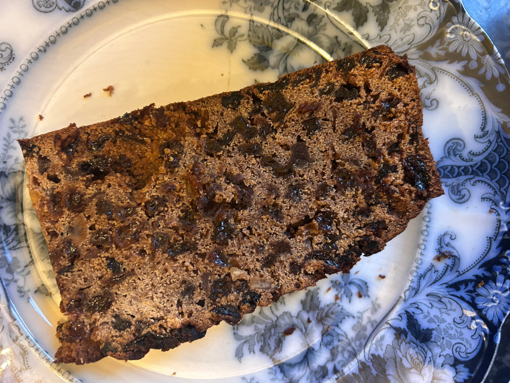 Slice of fruitcake resting on an antique-looking plate with a blue and white floral pattern. The cake is dark brown and densely packed with dried fruits, visible throughout the slice. The focus is sharp, highlighting the texture and ingredients of the cake.