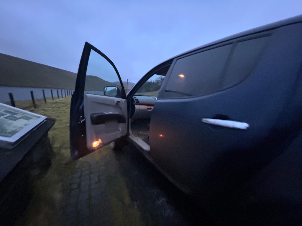 Dark-coloured pickup truck parked on a grassy area beside a long, low wall. The driver's side door is open, revealing the interior of the vehicle. The sky is a dusky blue, suggesting it is either early morning or late evening. A partially visible informational sign or map is lying on the ground near the truck. The overall mood is quiet and somewhat desolate.