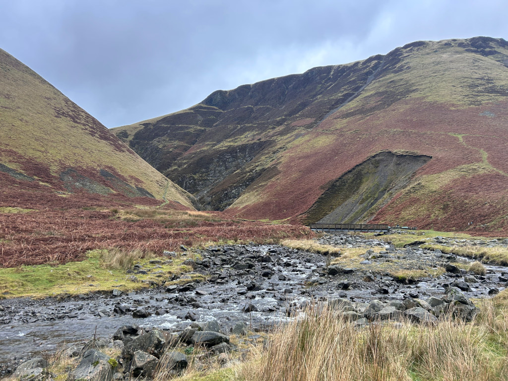 Tranquil landscape featuring a small stream meandering through a rocky valley. Two hillsides, one on either side of the stream, are covered in a mix of green grass and reddish-brown vegetation, possibly heather or bracken. A small, simple wooden footbridge spans the stream further up the valley. The overall tone is muted and peaceful, suggesting a remote, possibly mountainous, location. The sky is overcast, contributing to the calm atmosphere.