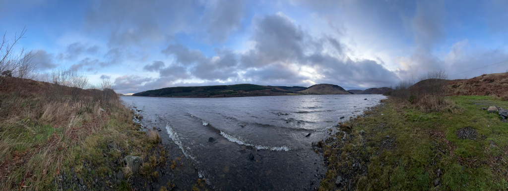 Panoramic view of a loch (lake) in Scotland. The water is relatively calm, with small waves lapping the shore. The banks of the loch are sparsely vegetated with scrub and grasses, typical of a temperate climate. In the background, hills rise gently, covered with what appears to be moorland vegetation. The sky is cloudy, but with some patches of blue visible. The overall impression is one of a peaceful and slightly melancholic, yet beautiful, natural landscape.