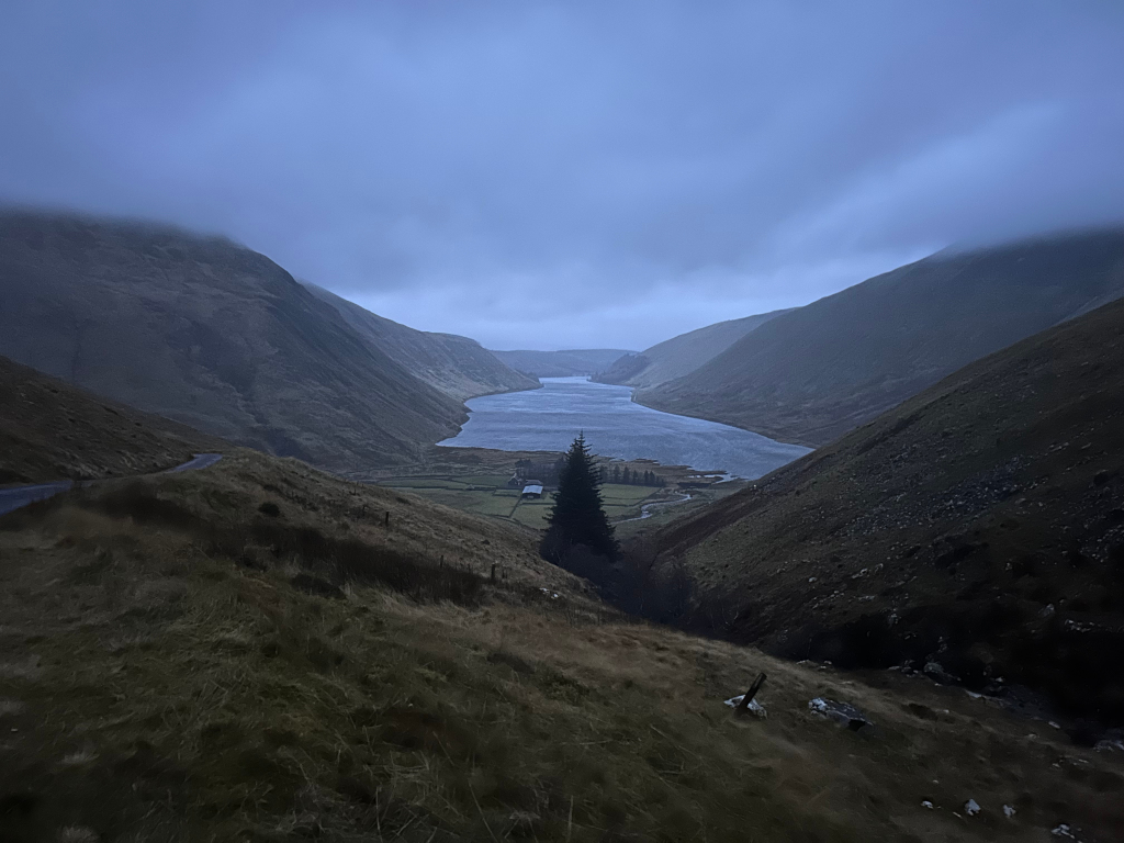 Serene yet somewhat sombre landscape.  A long, narrow lake or reservoir is nestled between two steep, dark green-brown mountains.  The sky is a muted, cloudy blue, adding to the overall quiet and somewhat moody atmosphere.  A single, dark evergreen tree stands out near a small structure at the base of one of the mountains, providing a tiny speck of contrast against the large scale of the landscape. A road winds around the mountainside in the foreground. The overall impression is one of isolation and the raw beauty of a remote landscape.