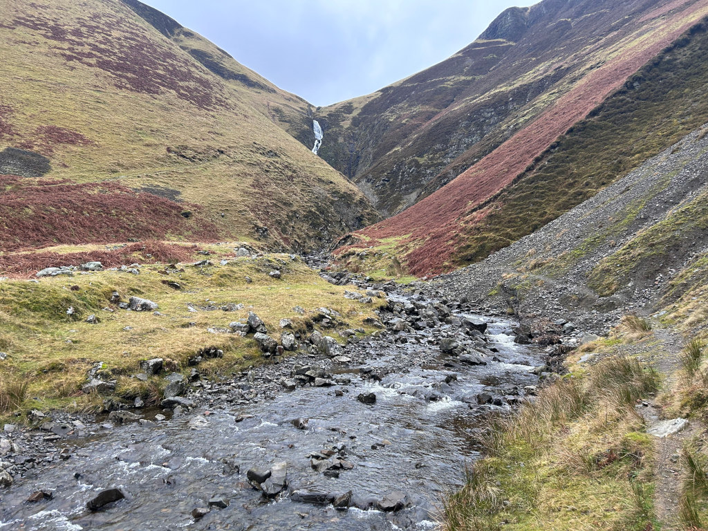 Mountain stream flowing through a rocky valley. The valley walls are steep, with patches of reddish-brown vegetation contrasting against the predominantly green and grey landscape. A small waterfall is visible high up on the valley wall.