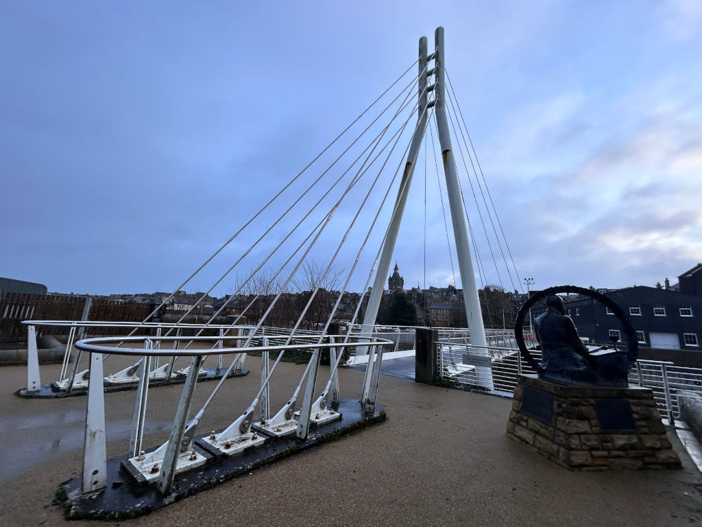 Modern, cable-stayed pedestrian bridge. In the foreground is a portion of the bridge's deck, featuring distinctive metallic railings and supporting structures. A bronze statue of a seated figure, possibly a historical figure or artist, is prominently placed on a stone pedestal near the bridge. In the background, a city scape is visible, suggesting an urban location. The sky is a muted blue-grey, indicating either dawn or dusk. The overall mood is calm and somewhat contemplative.