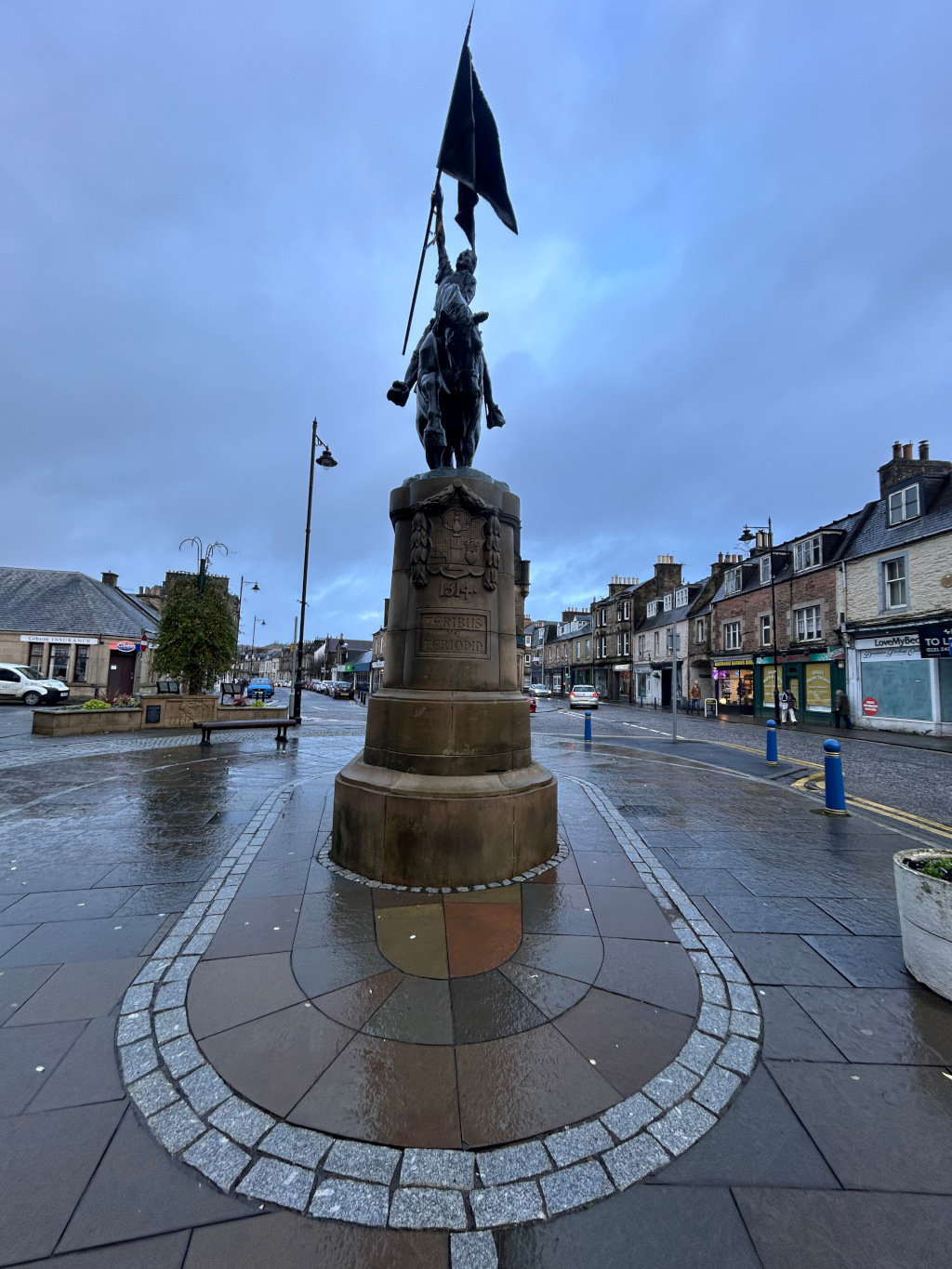 Statue of a mounted figure holding a flag atop a stone pedestal in a town square. The statue is dark-coloured bronze or similar material, and the flag is black. The square is wet, suggesting recent rain. Buildings typical of a small British town line the street behind the statue. The overall atmosphere is sombre and somewhat melancholic due to the dark colours, the overcast sky, and the wet paving stones. The inscription on the pedestal is partially visible.