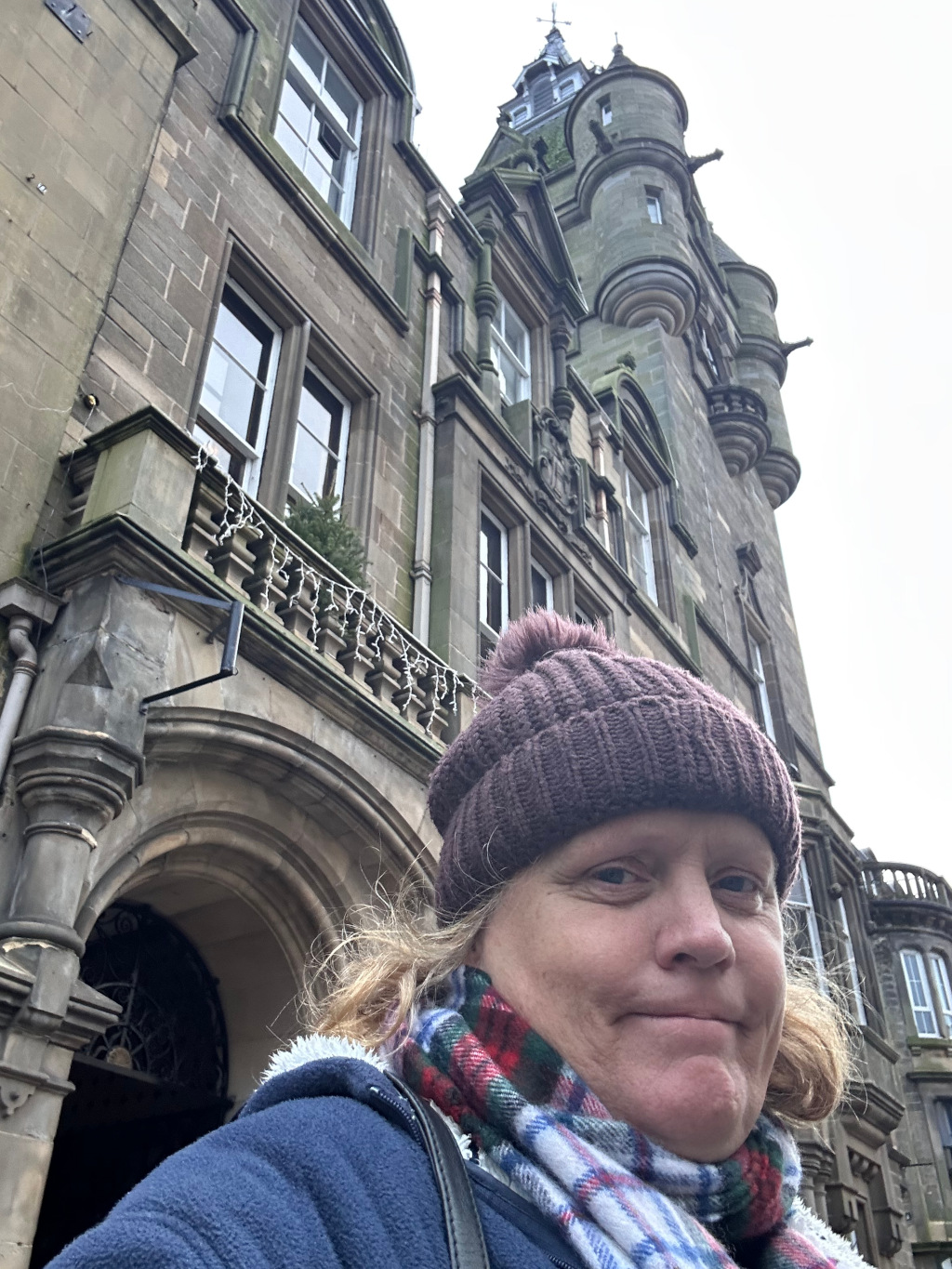 Leonie wearing a brown knit hat and a plaid scarf standing in front of a large, historic-looking building. The building is made of stone and has several stories, arched entrances, and towers. The woman appears to be taking a picture of herself and the building. The overall mood is casual and observational.