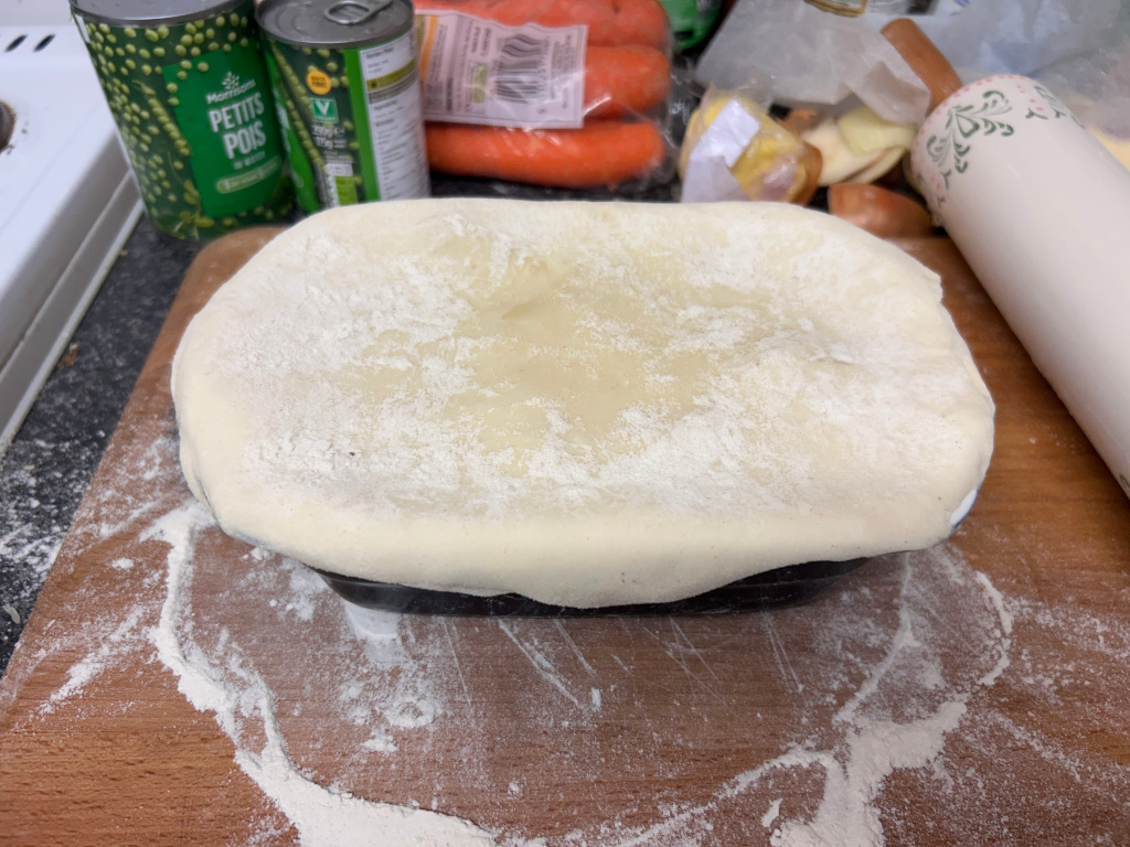 Rectangular baking dish filled with a savoury pie or similar baked dish. The dish is covered with a layer of uncooked pastry dough, lightly dusted with flour. In the background, there are cans of peas, carrots, and other food items, suggesting the preparation of a meal. A rolling pin is partially visible in the upper right corner. The overall impression is one of food preparation in a domestic kitchen setting.