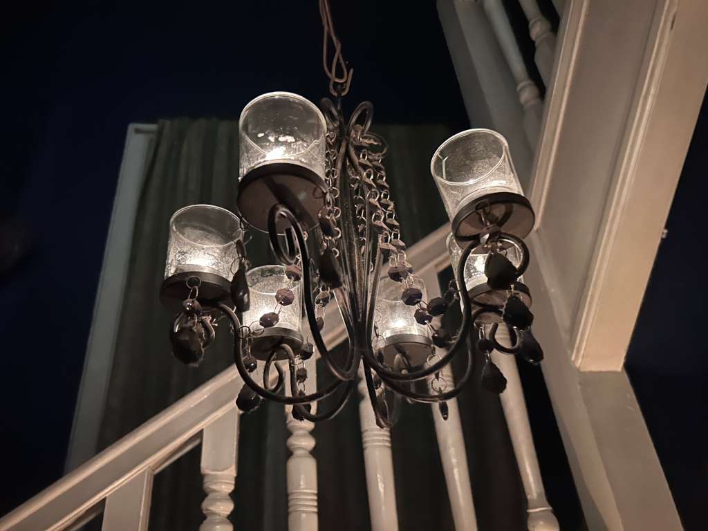 Dark-coloured, ornate chandelier with five candle holders, each containing a lit candle. The chandelier is suspended from the ceiling and hangs in front of a white staircase. The overall lighting is dim, creating a somewhat moody atmosphere.