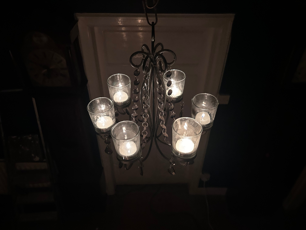 Dark room with a black metal candle chandelier hanging from the ceiling. Seven small glass candle holders are attached to the chandelier, each containing a lit tea light candle. The warm glow of the candles provides the only significant light source in the otherwise dark environment. The overall mood is dark, intimate, and somewhat mysterious.