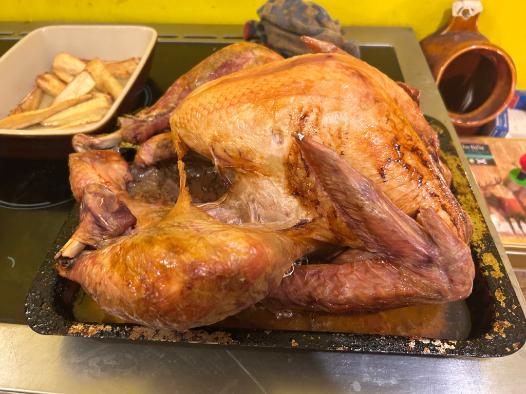 Roasted turkey on a dark metal roasting pan. The turkey is golden brown and glistening with juices. In the background, a separate dish contains roasted parsnips. The setting appears to be a kitchen counter-top with a yellow wall visible.