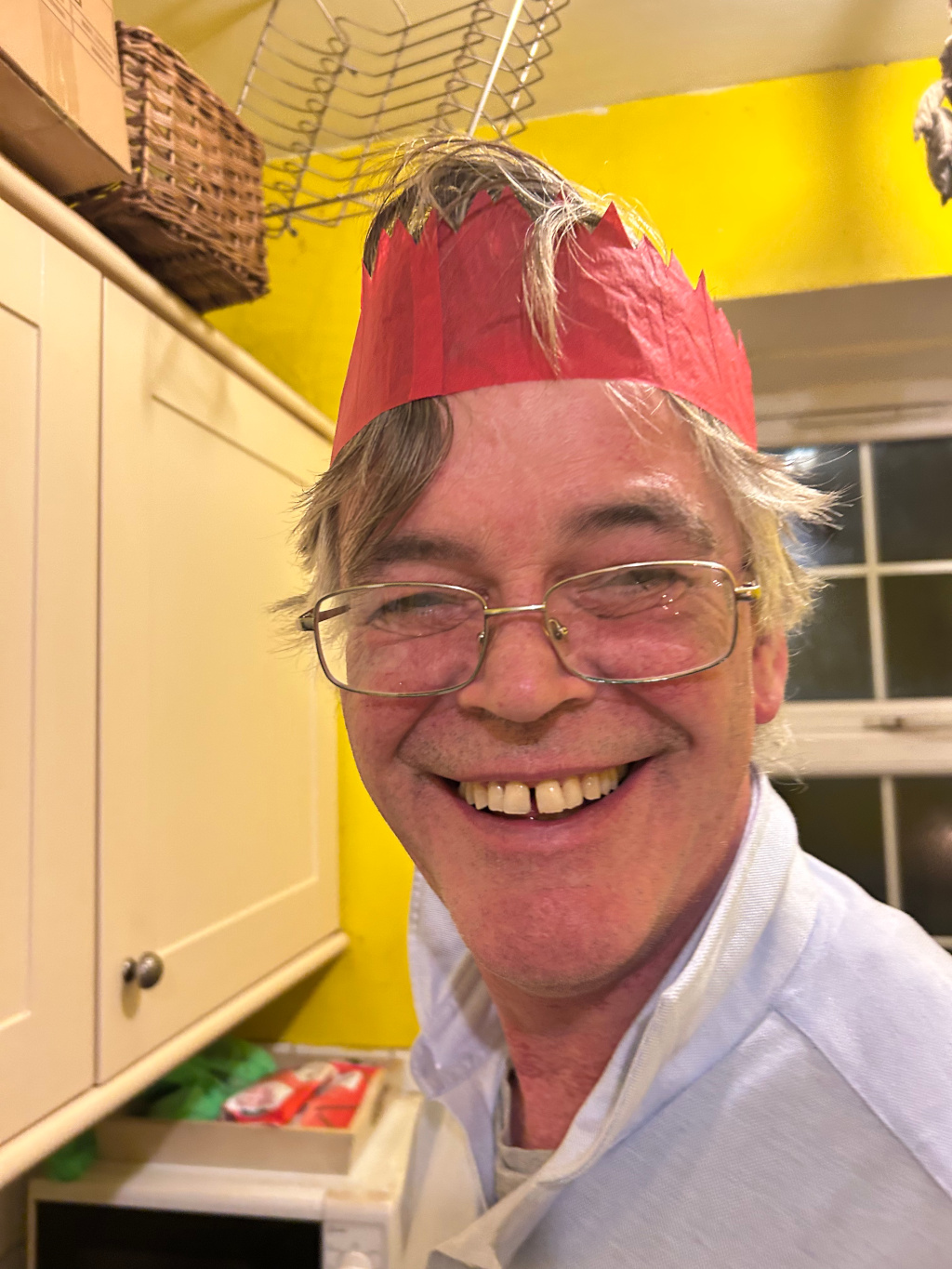 Close-up of a Charlie with glasses wearing a red paper Christmas crown. He is smiling broadly, showing his teeth, and his hair is slightly dishevelled. The background is a yellow wall with a kitchen cabinet and some kitchen utensils visible. The overall impression is one of festive cheer and informality.