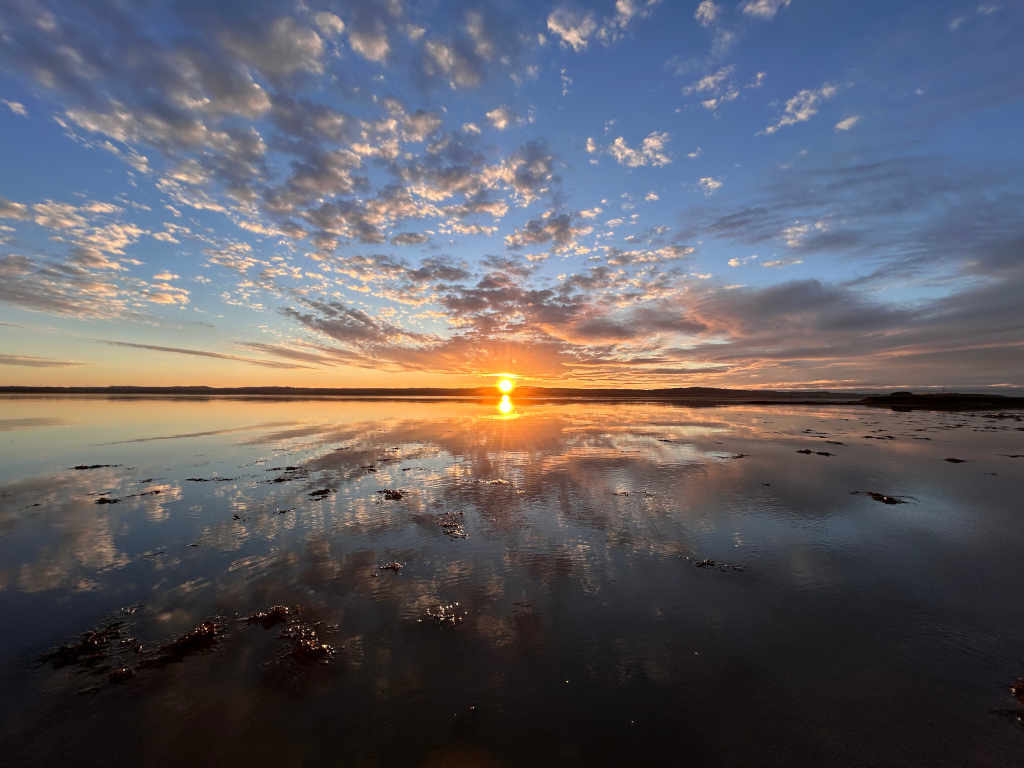 Serene sunset over a calm body of water. The sky is filled with a vibrant display of colours, ranging from warm oranges and yellows near the horizon to cooler blues and purples higher up. The clouds are scattered, creating a textured effect. The setting sun is visible on the horizon, its reflection mirrored perfectly in the still water. The water's surface is relatively calm, with patches of seaweed or other organic matter visible. The overall impression is one of peacefulness and natural beauty.