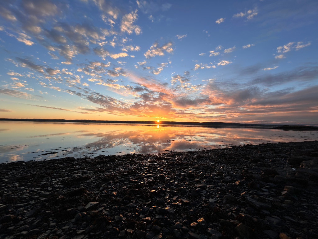Stunning sunset over a calm body of water. The sky is ablaze with vibrant colours of orange, pink, and purple, reflected beautifully in the still water. The foreground consists of a dark, rocky shoreline. The overall mood is serene and peaceful.