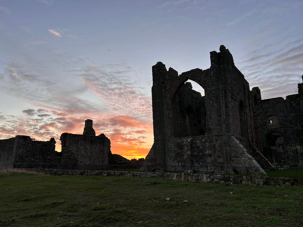 Silhouette of the ruins of an Abbey at sunset. The setting sun casts a warm orange glow on the sky behind the dark stone walls and towers of the abbey, creating a dramatic contrast. The image focuses on a prominent section of the ruins, with the rest of the structure visible in the background. The foreground is simple, a grassy field stretching towards the viewer.
