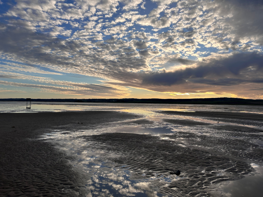 Tranquil coastal scene at either dawn or dusk. The expansive sky dominates, filled with a dramatic array of clouds – a mix of fluffy cumulus and darker, heavier formations. These clouds reflect beautifully in the shallow, tide-pool-like water covering a wide expanse of dark sand on a receding beach. A single, small, dark structure stands alone in the far distance near the waterline, adding a sense of solitude to the scene. The overall mood is serene and somewhat melancholic, emphasised by the soft light of the approaching or setting sun.
