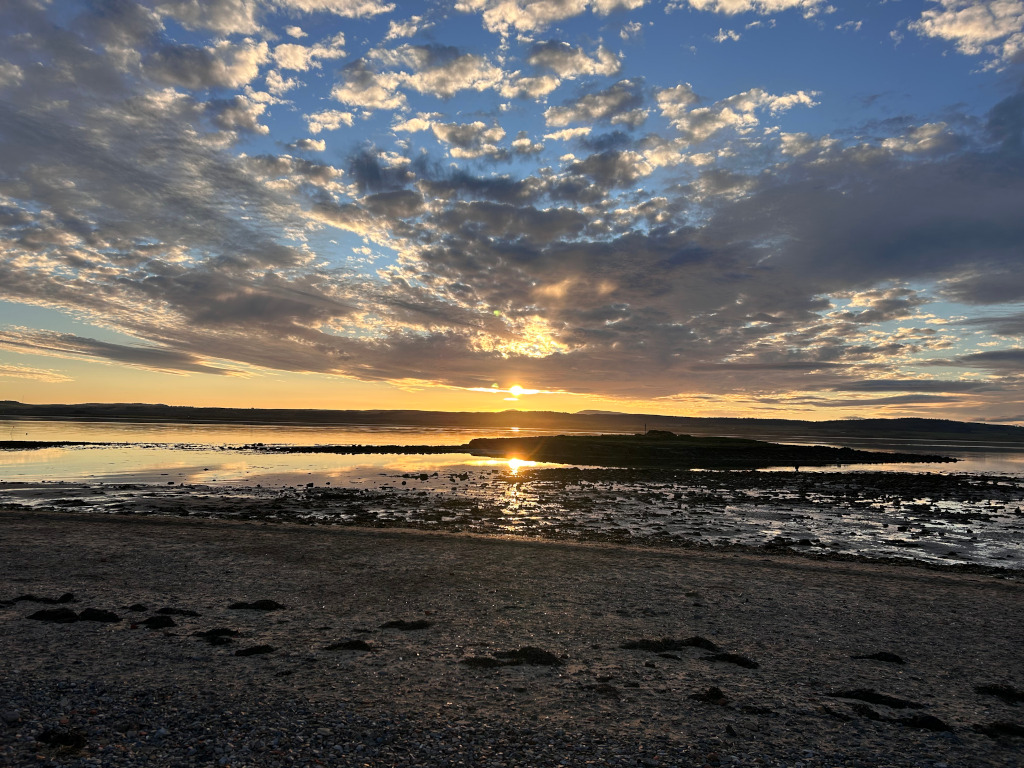 Serene sunset over a calm body of water, likely a bay or estuary. The sky is a breathtaking canvas of vibrant colours, with a mix of golden hues near the horizon, and cooler blues and greys higher up. The clouds are scattered, allowing the sun to shine through, casting a warm golden light on the water's surface and creating a reflection of the sunset. The foreground shows a pebbled beach, with small dark patches suggesting seaweed or rocks left by the receding tide. The overall mood is peaceful and tranquil, evoking a sense of calm and the beauty of nature.