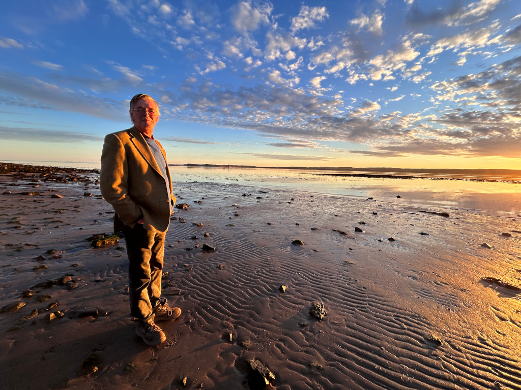 Charlie standing on a beach at sunset. He's wearing a tan blazer and khaki pants, and his pose suggests a contemplative or reflective mood. The setting sun casts a warm golden light across the wet sand, which shows ripple patterns left by the receding tide. The sky is a vibrant blend of blues, oranges, and clouds. The overall impression is one of serenity and perhaps introspection.