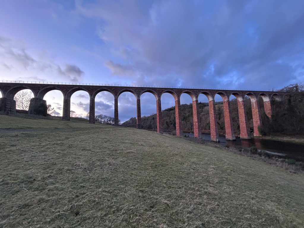 A long, red brick viaduct spanning a river. The viaduct has many arches and runs horizontally across most of the image. It's situated in a landscape of gently sloping grassland, with trees visible in the background, on a partly cloudy, dusk-like sky. The overall impression is one of serene, quiet grandeur.