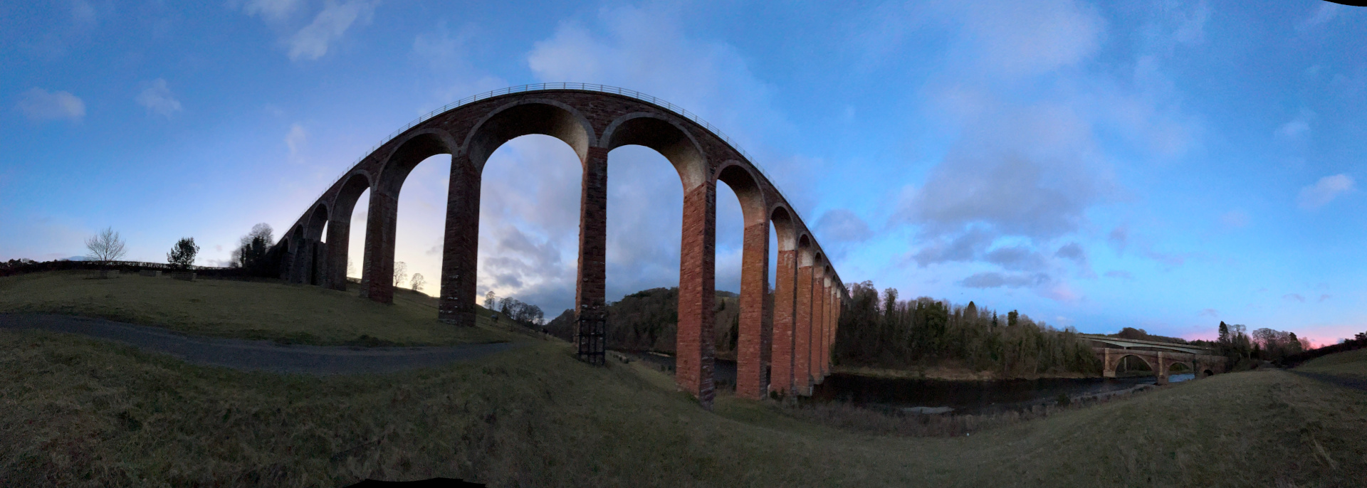 Panoramic view of a large brick viaduct curving gently across the landscape. The viaduct is composed of numerous arches, and it spans a river. The time of day is twilight; the sky is a mix of blue and pinkish hues, indicating either dawn or dusk. Grassy hills and a line of trees are visible in the foreground and background, respectively. The overall impression is one of tranquil beauty and a sense of scale, highlighting the impressive engineering of the viaduct against the serene natural setting.