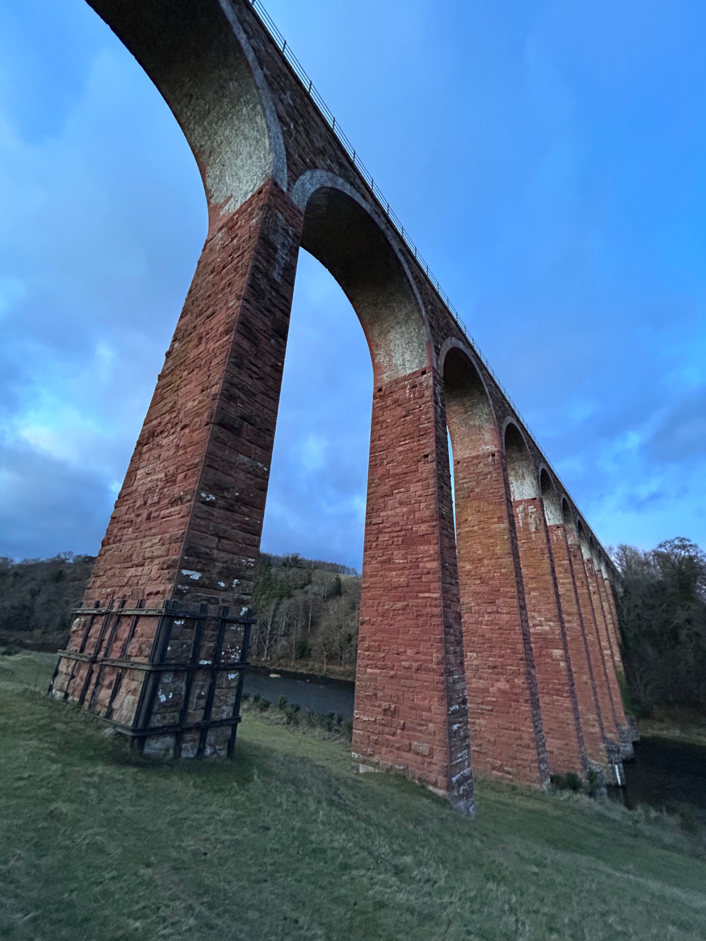 Low-angle view of a long, red-brick viaduct against a twilight sky. The viaduct consists of multiple tall, arched structures, and extends into the distance. A body of water and some trees are visible in the background. The overall impression is one of age, grandeur, and perhaps a sense of quiet solitude.