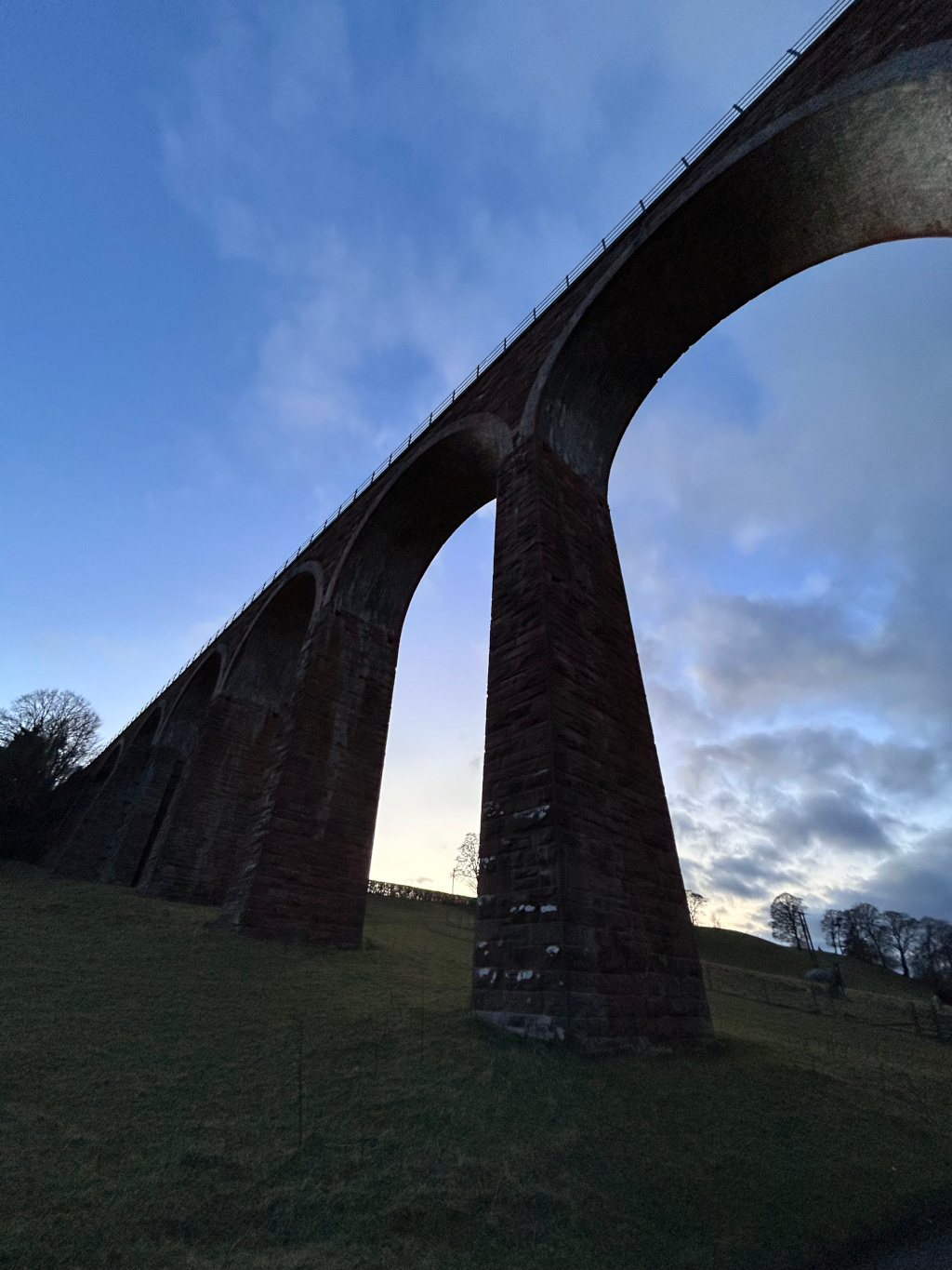 Low-angle view of a large stone viaduct against a twilight sky. The viaduct is composed of several tall, arched structures, and appears to be made of dark reddish-brown stone. The sky is a mix of blue and clouds, suggesting dusk. The foreground shows a gently sloping grassy hill. The overall impression is one of grandeur and a sense of scale, emphasising the height and length of the structure.