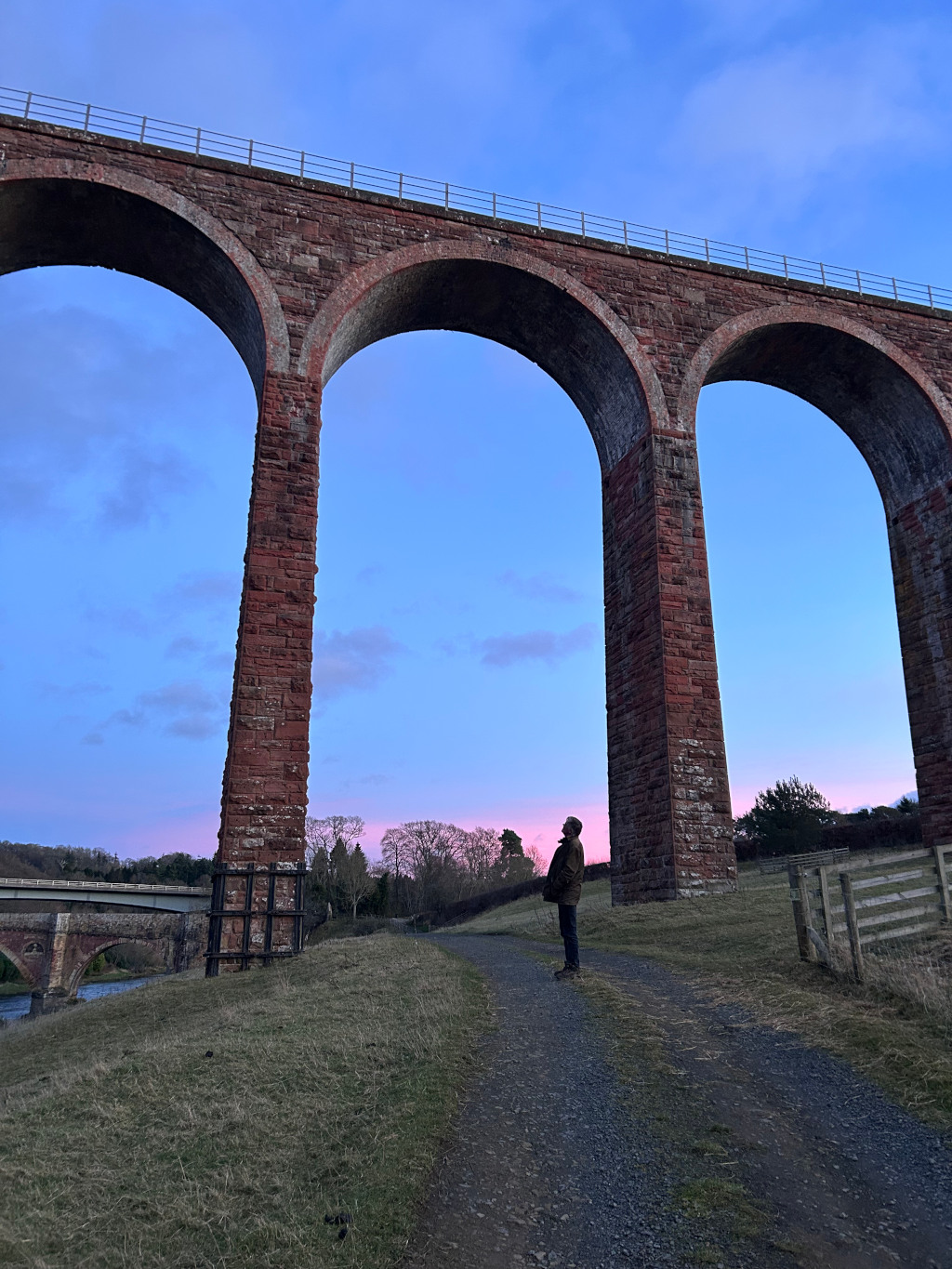 Standing on a gravel path, gazing upwards at a large brick viaduct against a twilight sky. The viaduct's imposing structure dominates the frame, with its arches creating a strong visual element. The soft, pastel colours of the sky at dusk add a serene and contemplative mood to the scene. A smaller bridge is visible in the distance across a river. The overall impression is one of scale, solitude, and the beauty of a natural and man-made landscape merging.