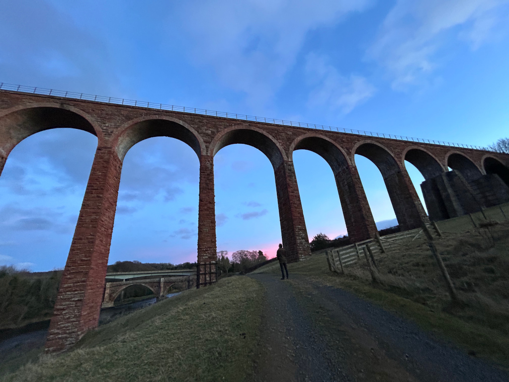 Low-angle view of a large brick viaduct stretching across the frame under a twilight sky.  A single person stands on a path leading away from the viaduct, providing a sense of scale. In the distance, a smaller bridge is visible over a body of water. The overall mood is serene and somewhat melancholic, with the soft colors of the twilight sky contrasting with the imposing structure of the viaduct.