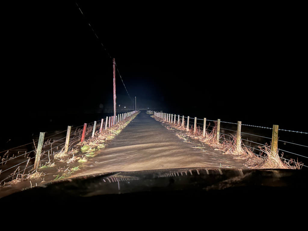 Nighttime view from inside a vehicle driving down a long, straight dirt road. The road is bordered by a wire fence with wooden posts, and the surrounding area appears to be flooded or very wet, with vegetation visible along the roadside. The only light source is the vehicle's headlights, which illuminate the road ahead and create a sense of depth and isolation. The dark sky and lack of other artificial light contribute to a somewhat ominous or lonely atmosphere.
