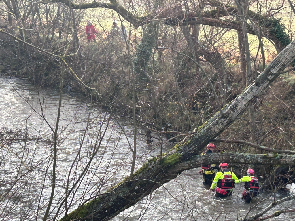 River scene where three rescue workers in bright yellow and red life vests are wading in a shallow, somewhat murky river. They seem to be engaged in a rescue operation, possibly searching for something or someone. In the background, there are bare winter trees and a few figures in the distance, appearing to be onlookers or additional emergency personnel. The overall mood is serious and slightly tense, indicative of an ongoing emergency situation.