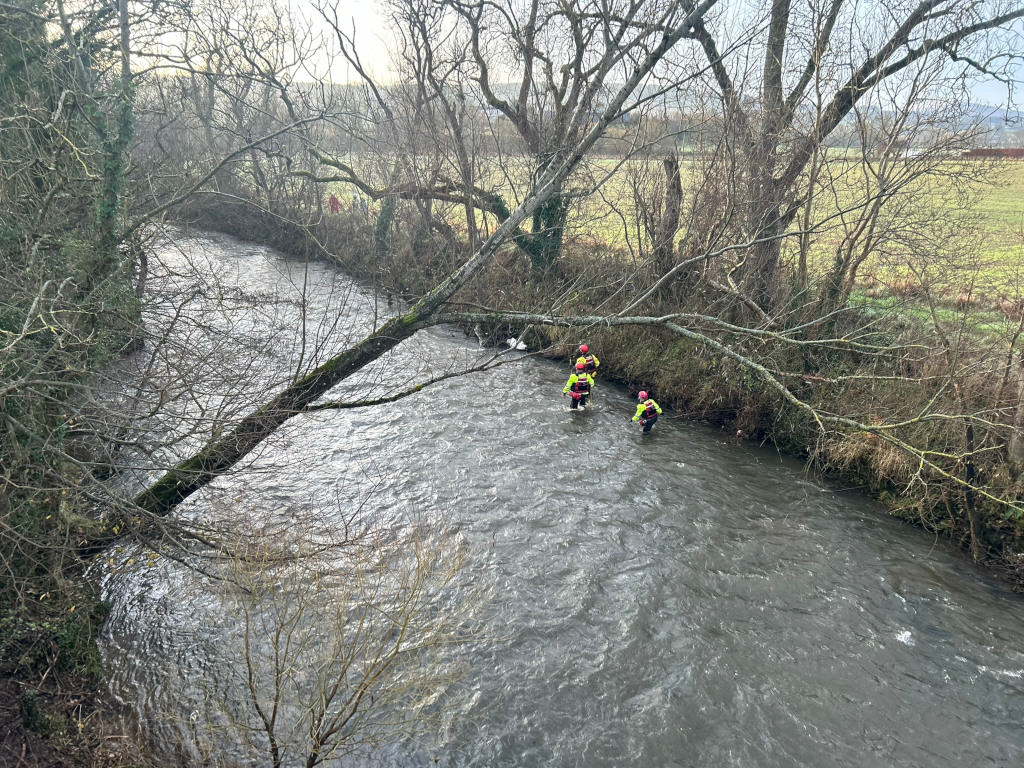 Three figures in bright yellow and dark clothing wading through a shallow, flowing river. They appear to be emergency personnel, possibly rescuers or search and rescue team members, given their attire and the context of their activity in the water. The river flows through a natural landscape with leafless trees lining its banks. The overall mood is somewhat somber and suggestive of an ongoing operation or search.