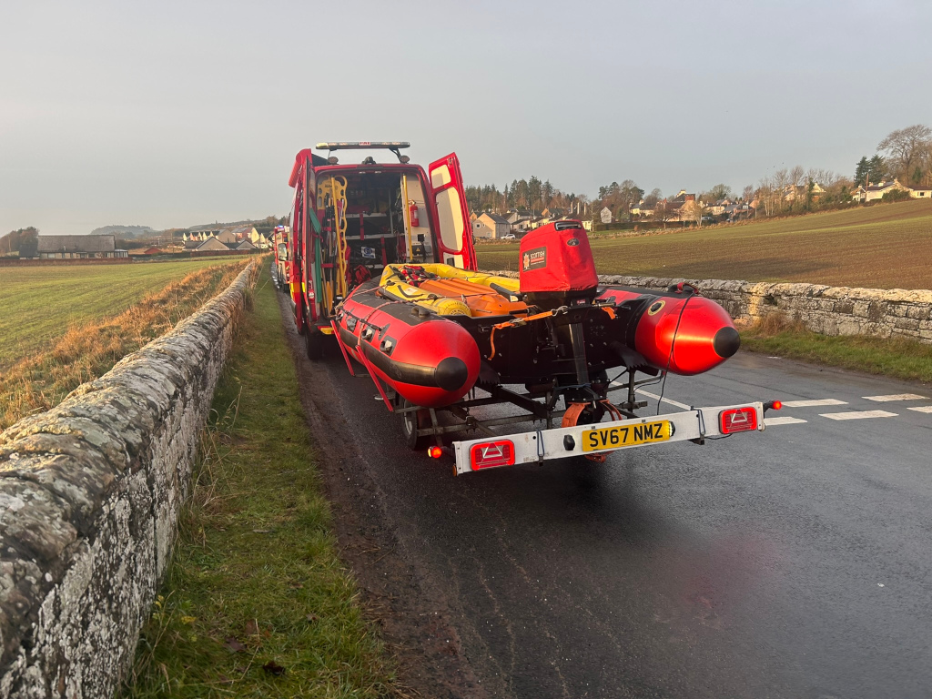 Red emergency response vehicle, with its rear doors open, parked on a road next to a low stone wall. A red inflatable boat on a trailer is attached to the back. The scene is rural, with fields and a small village visible in the background. The overall impression is that emergency services are responding to an incident in a rural location.
