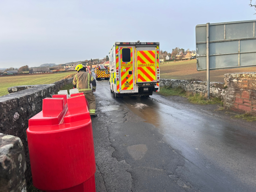 Fireman standing near a red barrier on a bridge. In the background, an ambulance and another emergency vehicle are visible. The scene seems to be post-incident, possibly at the site of an accident or emergency situation in a rural area. The overall mood is serious and suggests an emergency response is underway.