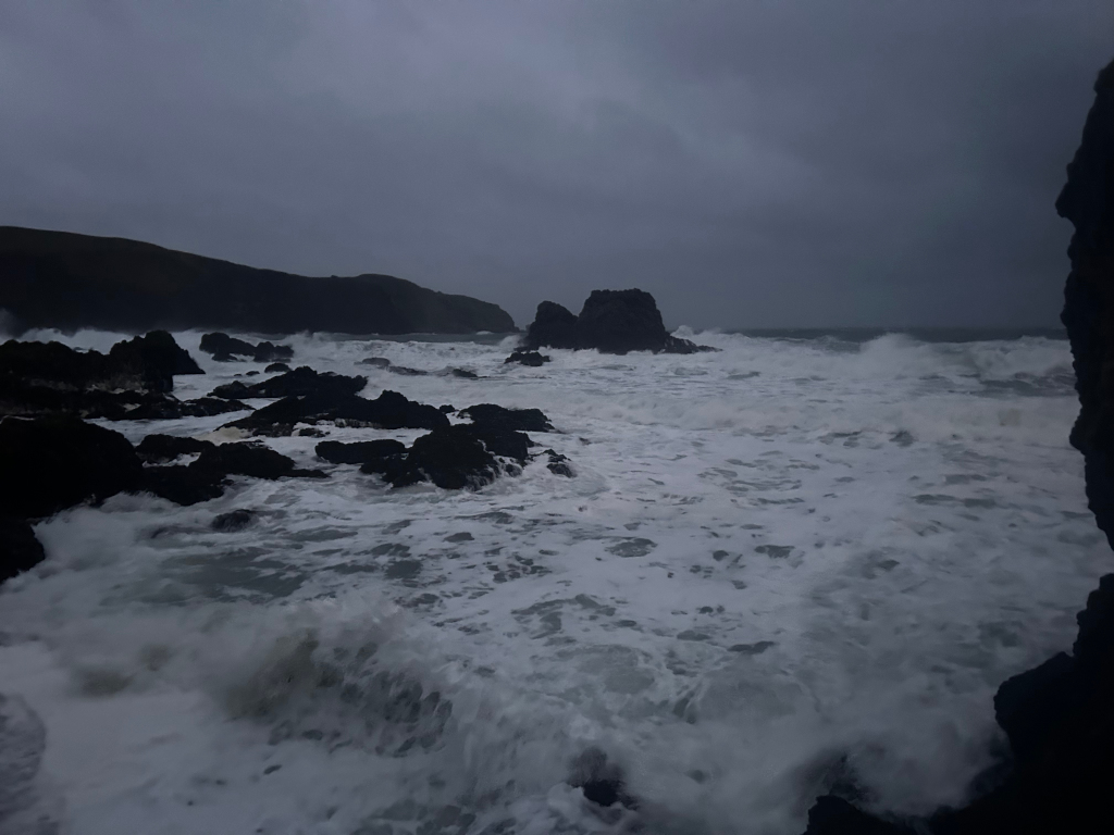 Dramatic seascape under a stormy sky. Rough waves crash against dark, rocky shoreline. The overall mood is one of wildness and power, with a muted colour palette emphasising the grey sky and churning white water. A sense of isolation is conveyed by the vast expanse of ocean and the rugged, seemingly uninhabited coastline.