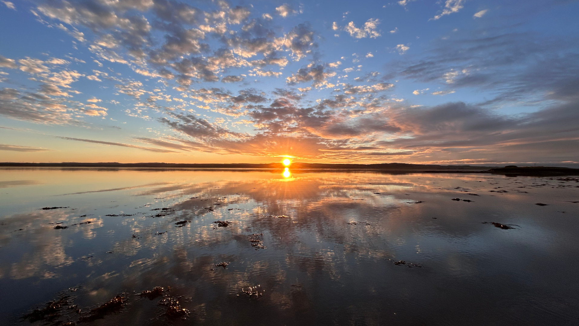 Breathtaking sunset over a calm body of water. The sun, a vibrant orange-yellow, sits low on the horizon, its light casting a warm glow across the sky and reflecting beautifully on the still water. The sky is a stunning canvas of colours, with hues of orange, pink, and purple blending seamlessly into a clear blue. Patches of seaweed are visible on the water's surface, adding texture and depth to the scene. The overall atmosphere is serene and peaceful, evoking a sense of tranquillity and awe.