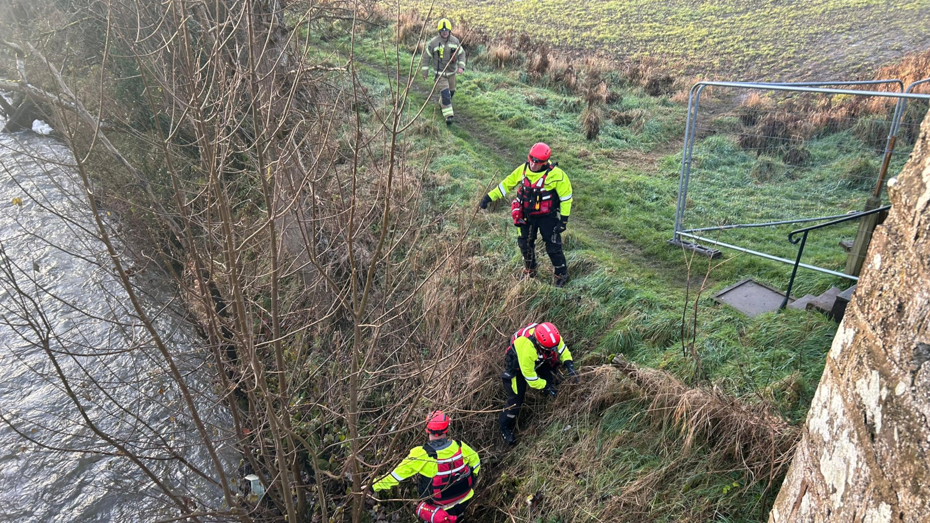 High-angle view of four emergency personnel near a riverbank. Three are wearing bright yellow and red rescue gear and are actively searching along the riverbank, which is overgrown with vegetation. One person, further up a path, appears to be in firefighting gear. The scene suggests a search and rescue operation.
